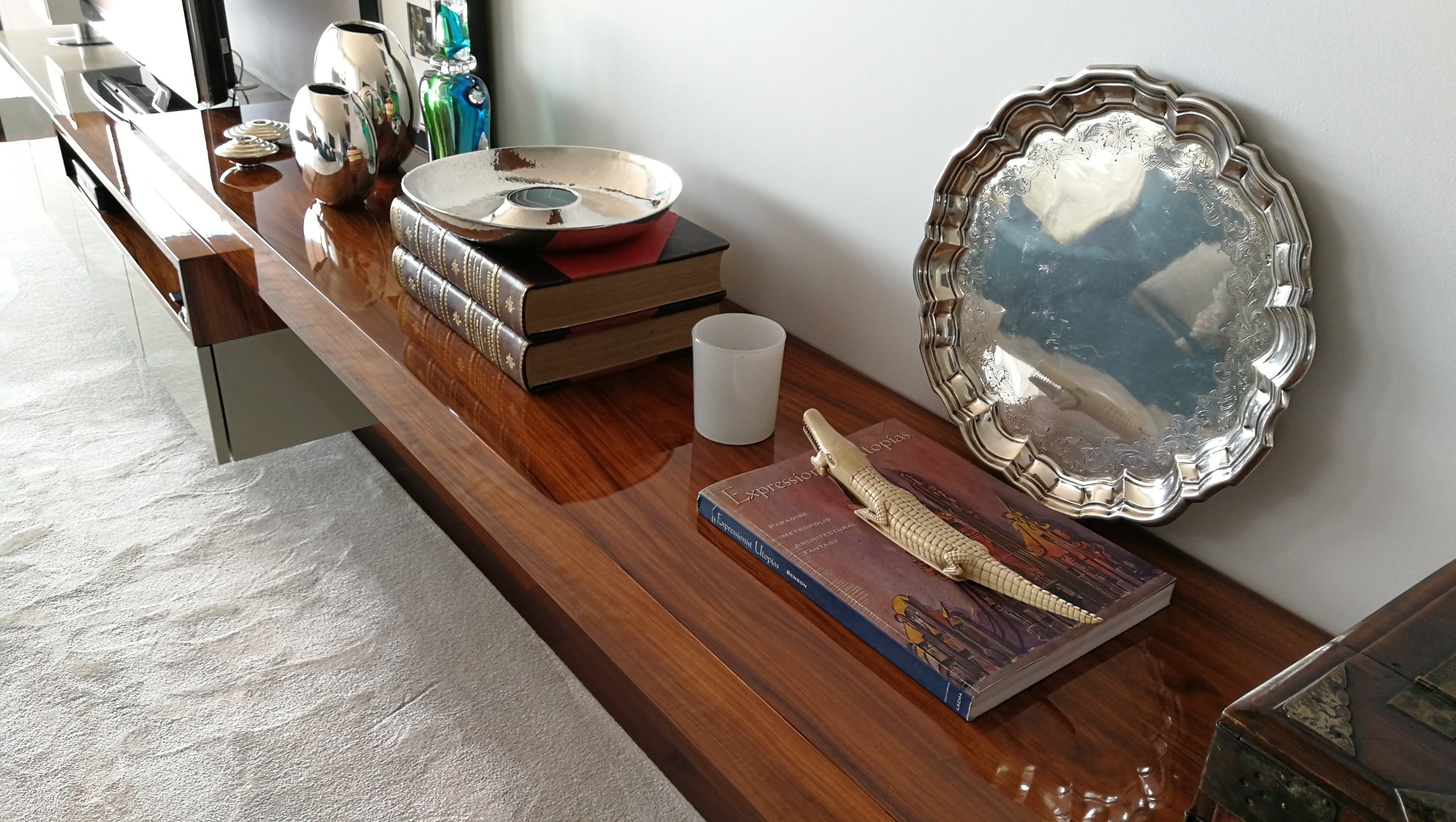 Aparador suspenso em madeira de nogueira com acabamento alto brilho, decorado com jarras prateadas e objetos decorativos