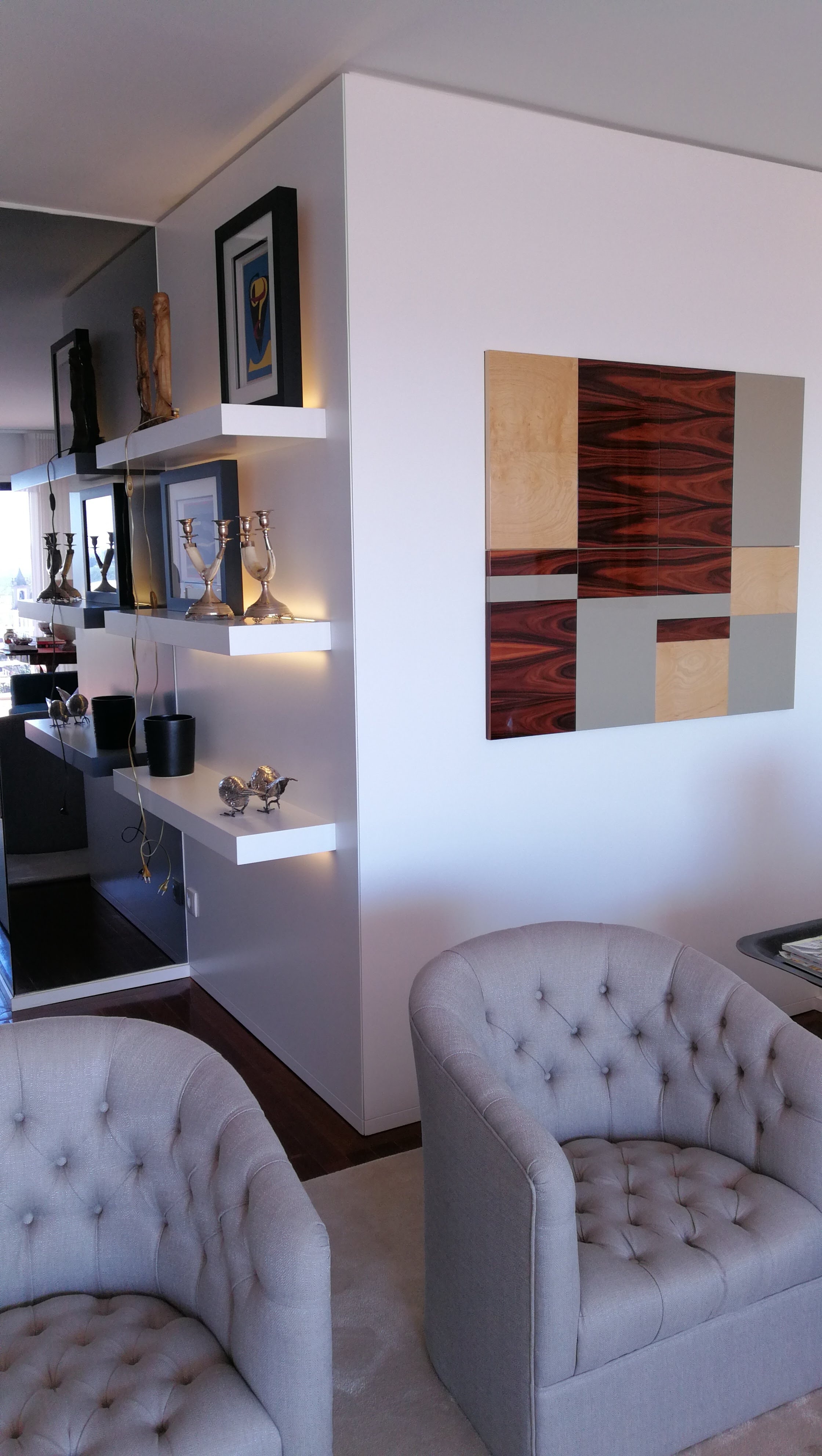 Zona de estar com poltronas capitone cinza, prateleiras flutuantes iluminadas e painel decorativo em madeira