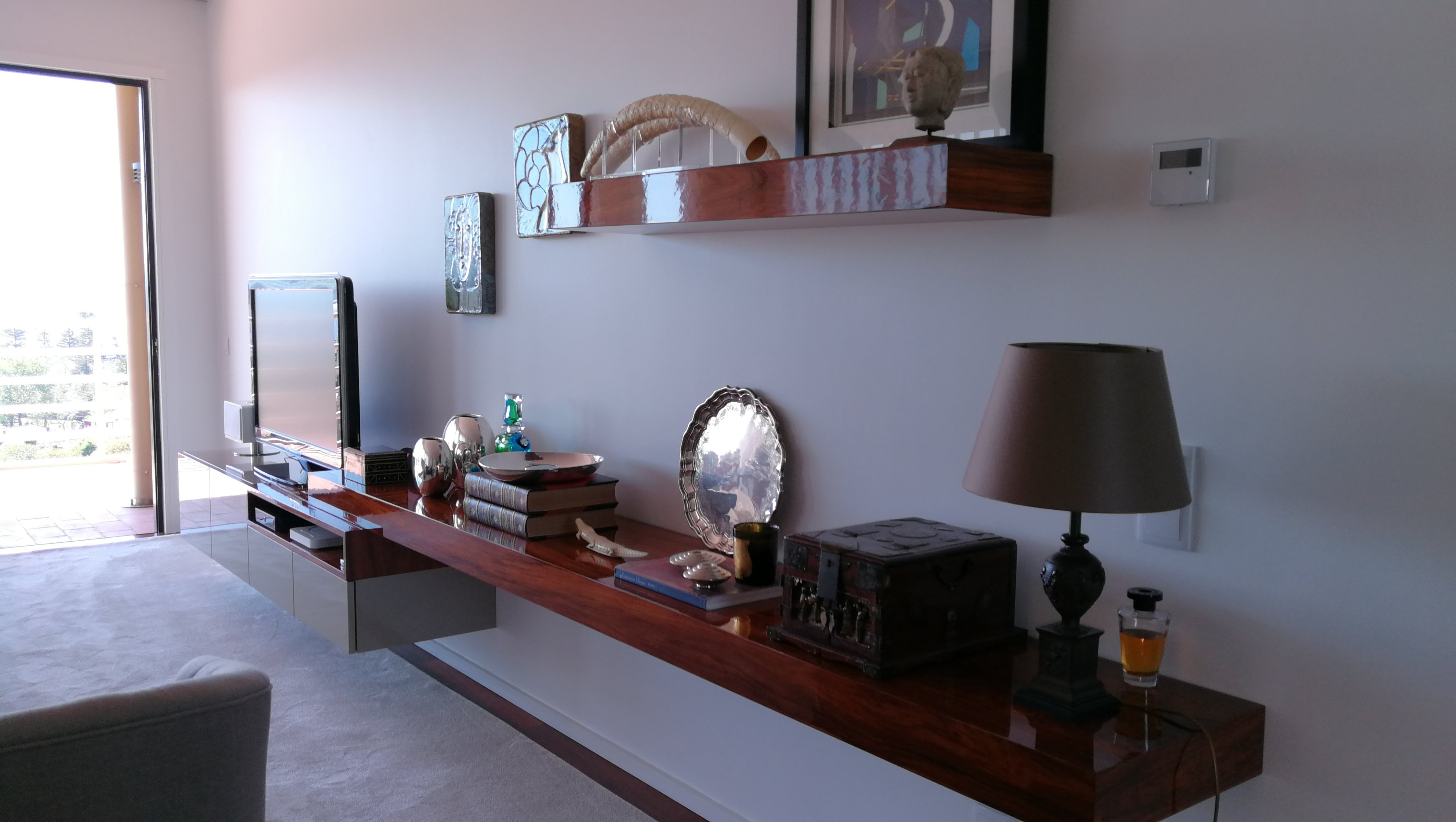 Vista geral da sala com movel suspenso em nogueira alto brilho, prateleira com esculturas, candeeiro e porta de varanda