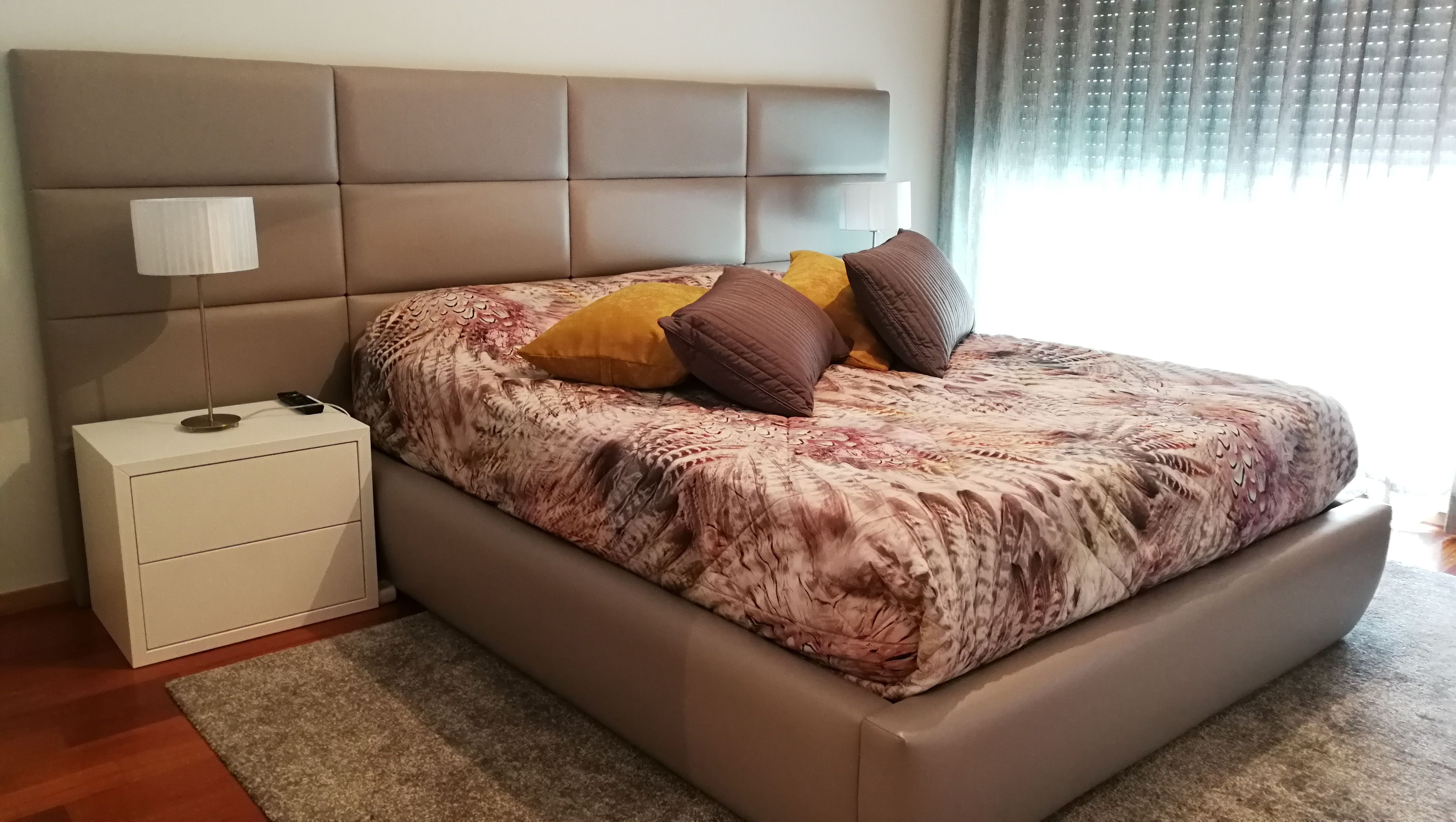 Quarto com cama estofada em pele cinza-taupe, mesas de cabeceira brancas e roupa de cama em tons rosa e castanho