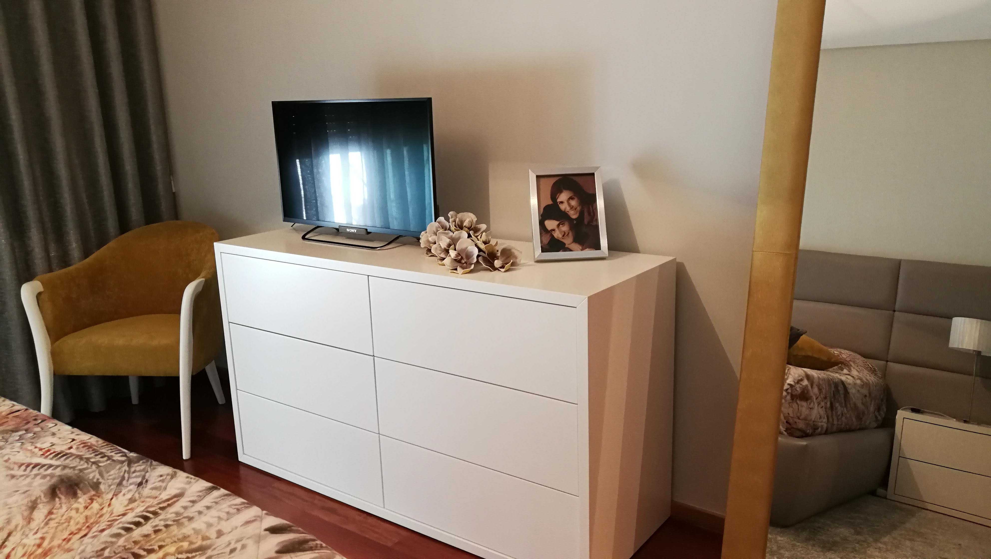 Quarto com comoda lacada branca, televisao, poltrona mostarda e cabeceira estofada cinza