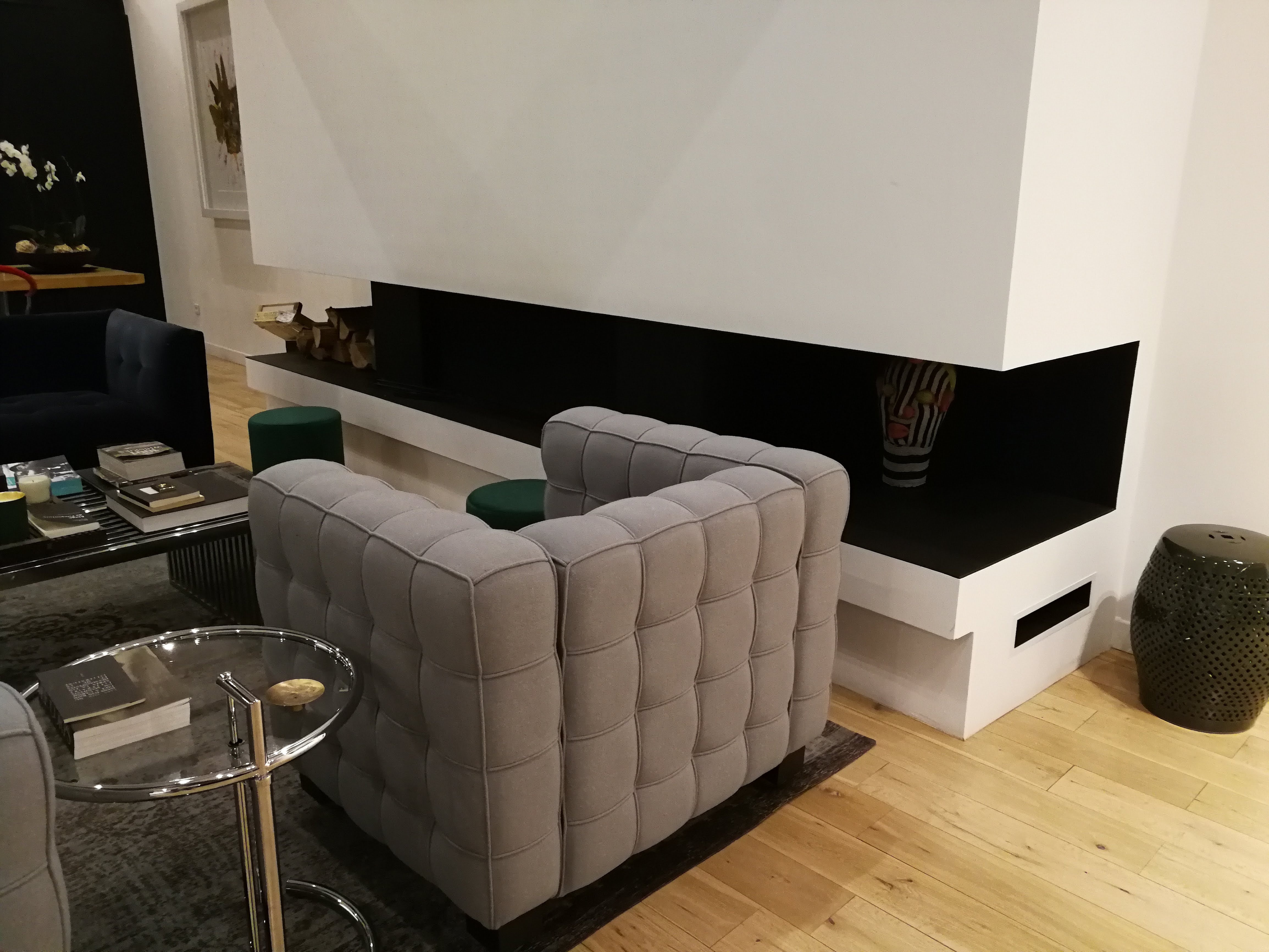 Sala com poltrona capitone cinza, lareira moderna embutida, mesa cromada e pufes verdes