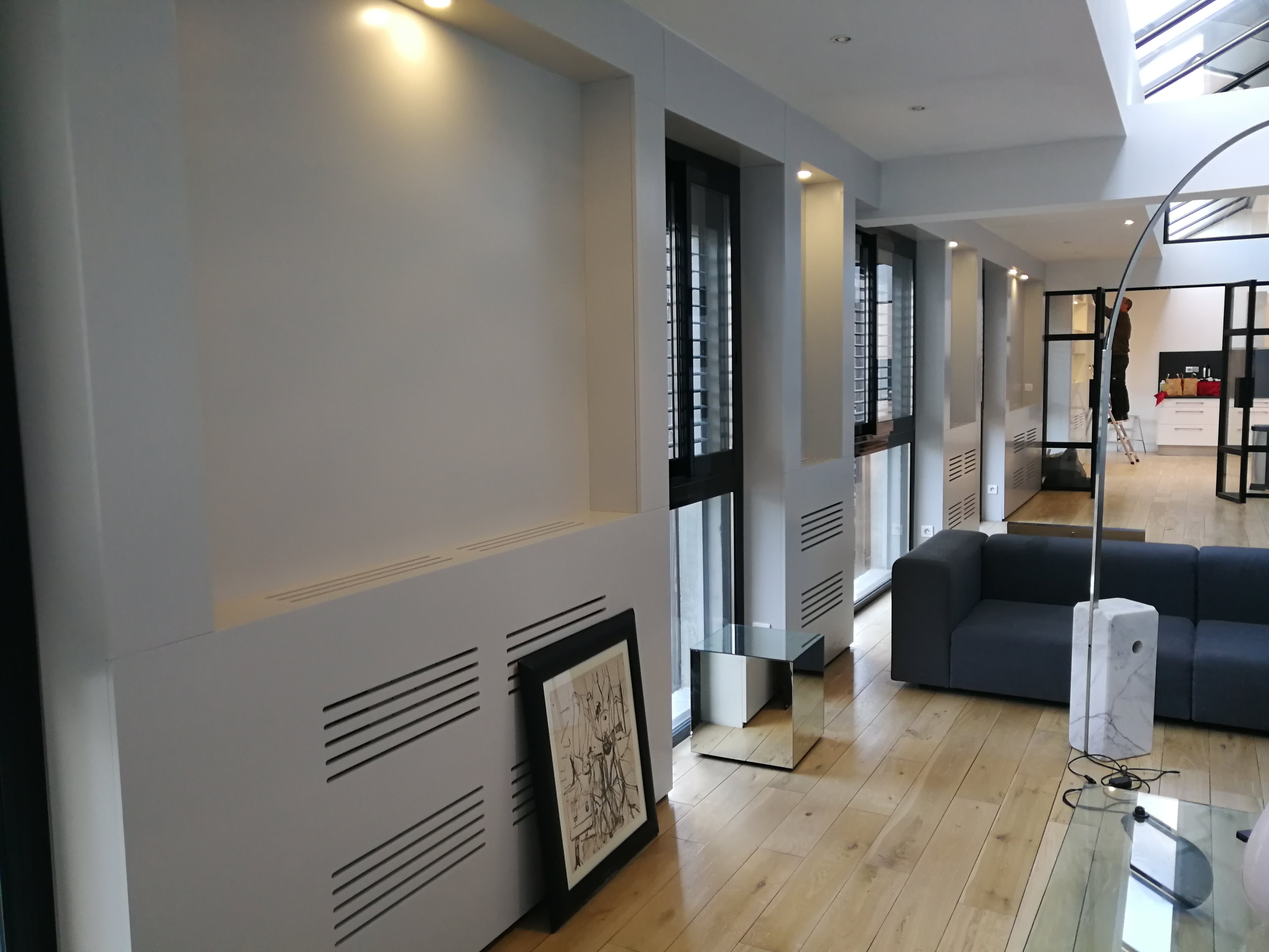 Sala com paineis brancos entre janelas altas, sofa modular cinza, candeeiro arco e mesa espelhada