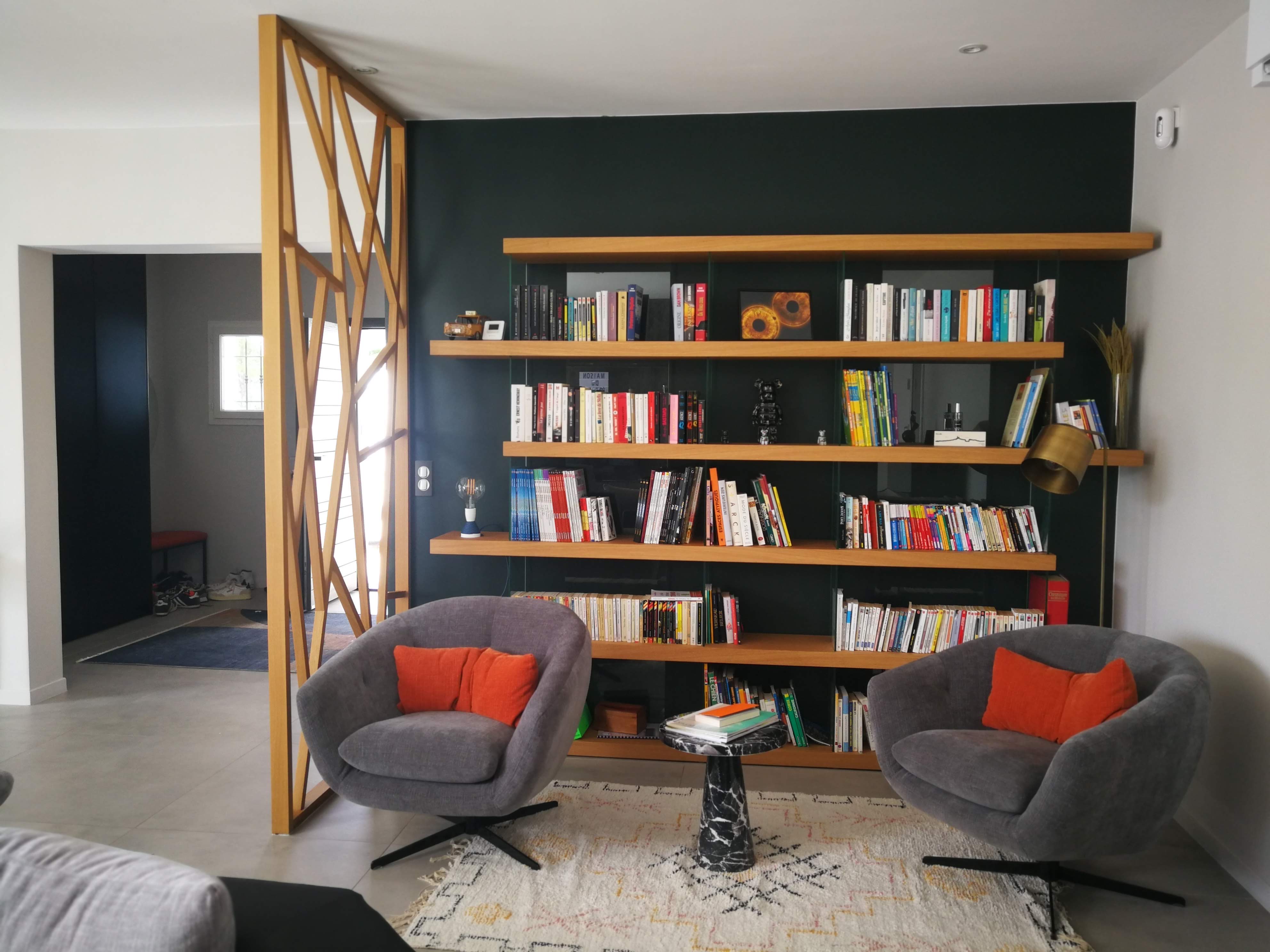 Zona de leitura com estante em carvalho sobre parede verde, poltronas cinza com almofadas laranja e divisoria em madeira