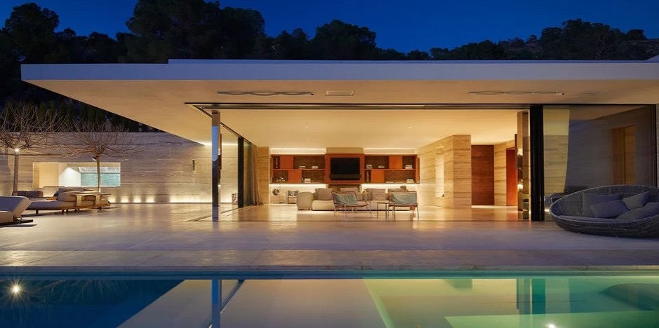 Vista noturna da villa com piscina infinita, sala aberta com paredes de vidro e iluminacao quente