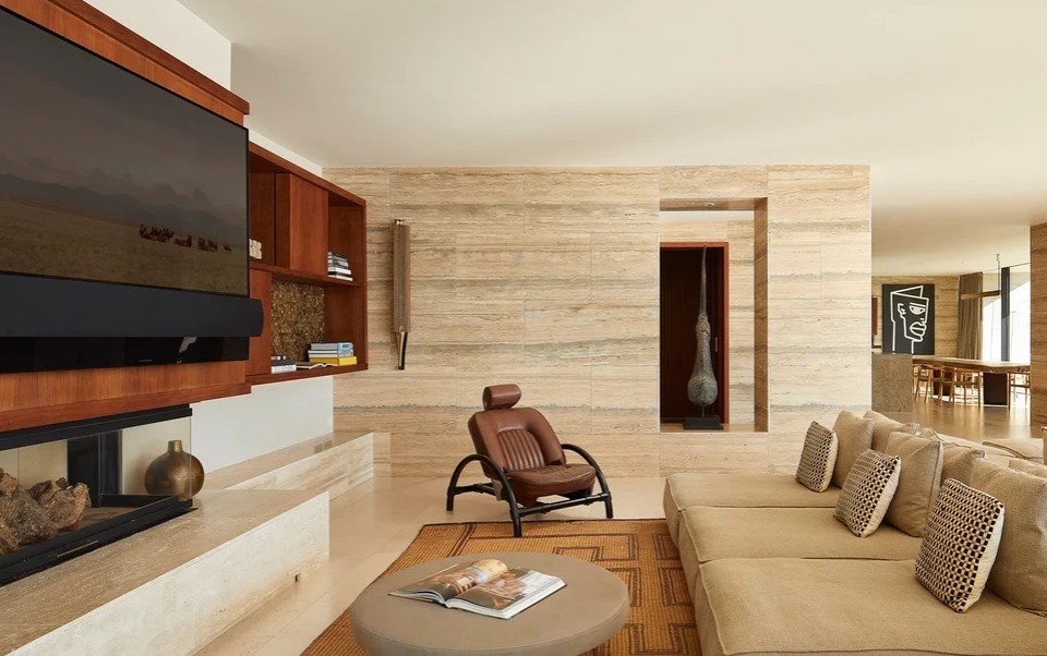 Sala com sofa bege, poltrona em couro, lareira embutida, movel TV em madeira e parede em travertino