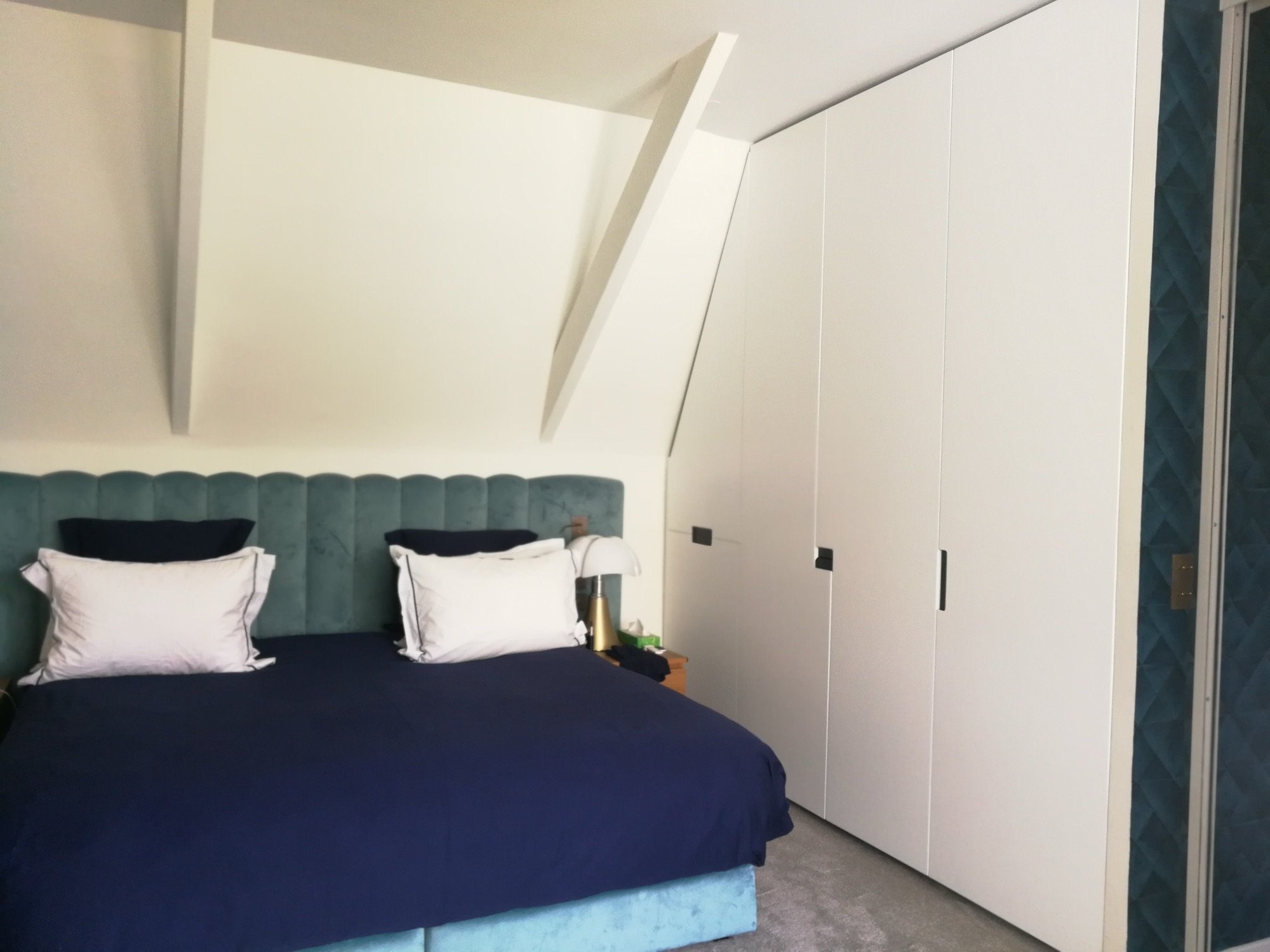 Suite em sotao com cabeceira estofada verde-azulada, armario embutido branco e papel de parede geometrico