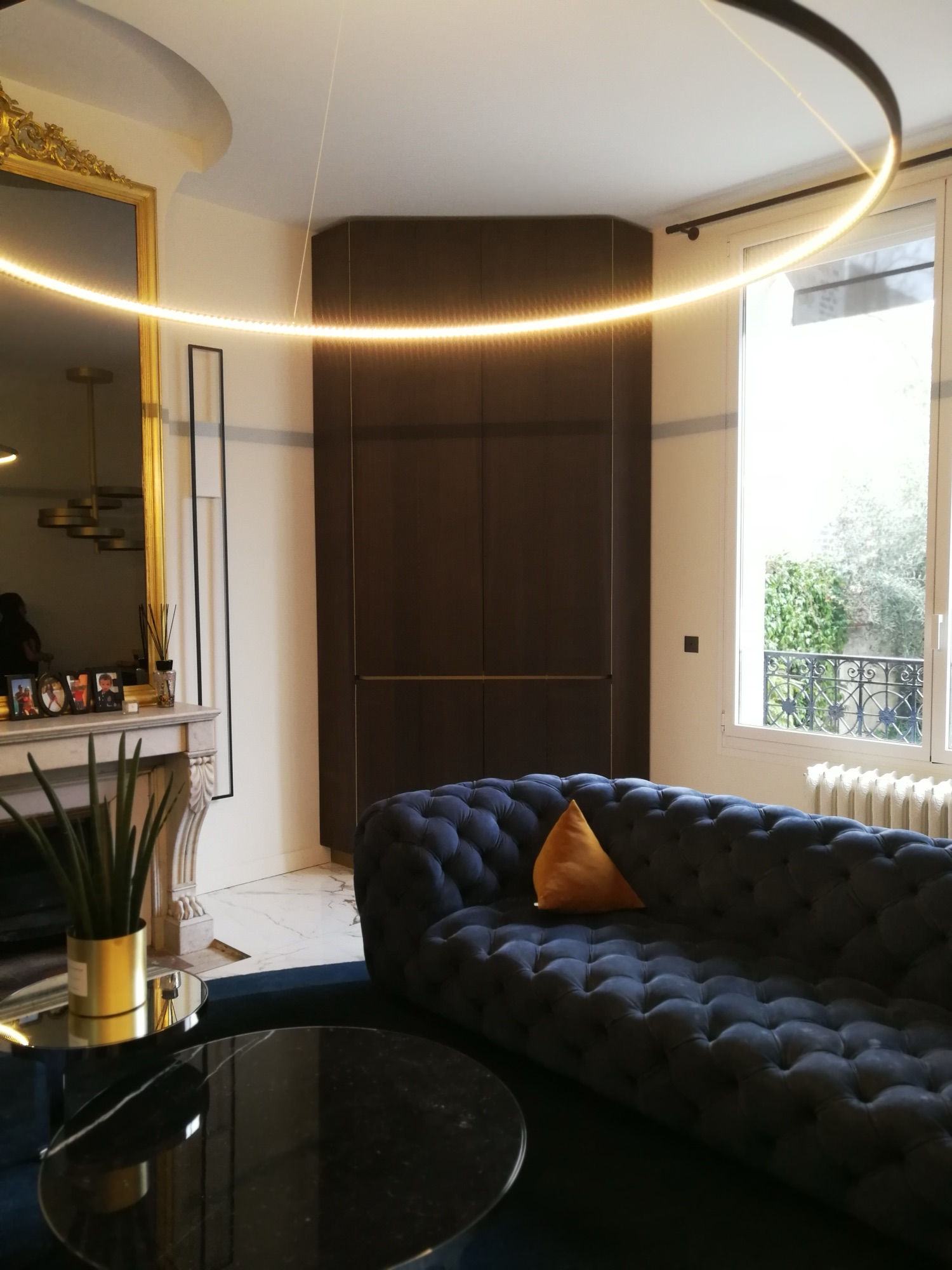 Sala com sofa Chesterfield azul-marinho, armario em madeira escura, lareira em marmore e candeeiro circular