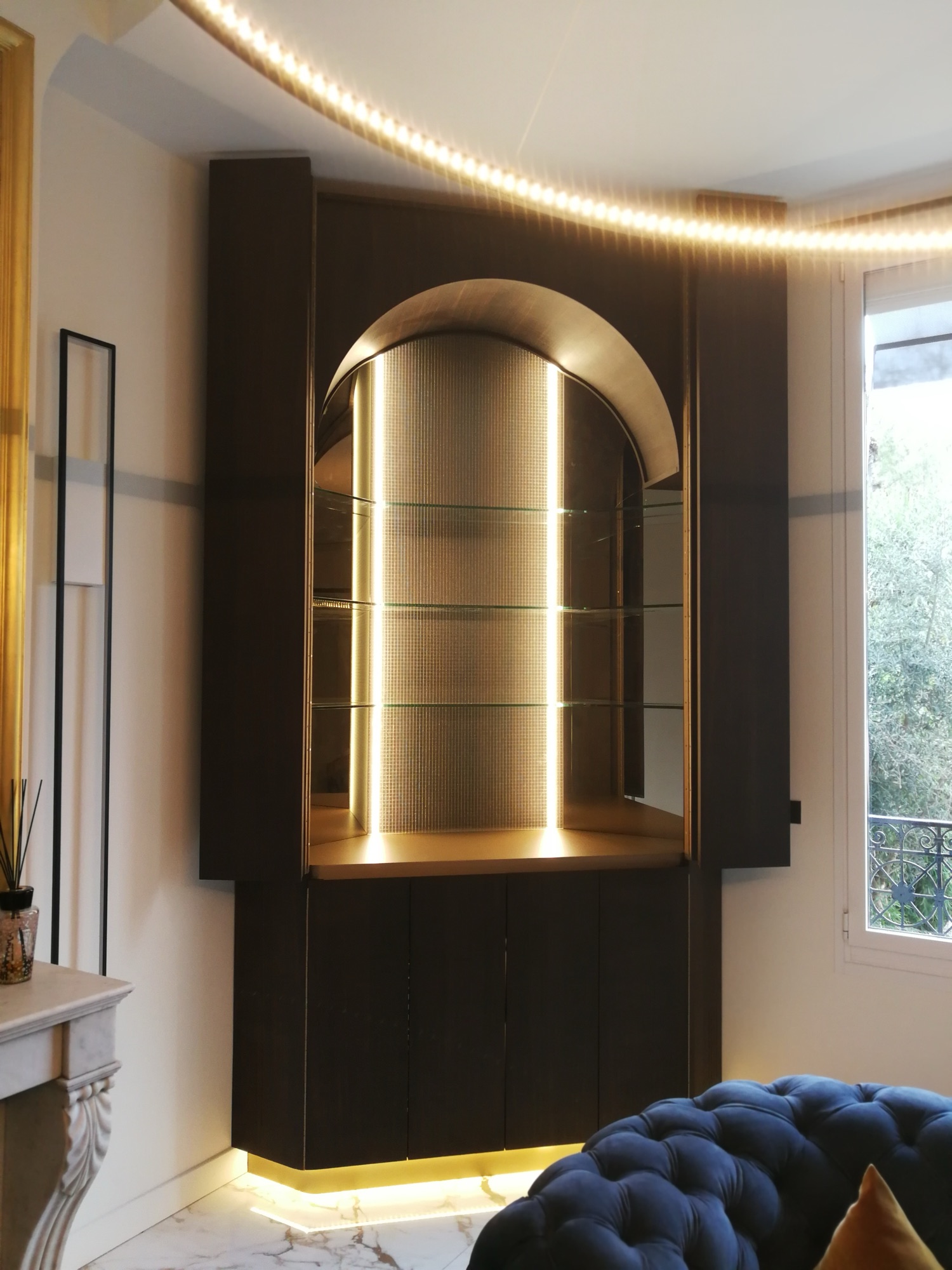 Vitrina/bar em arco dourado com prateleiras de vidro iluminadas e fundo em malha metalica dourada