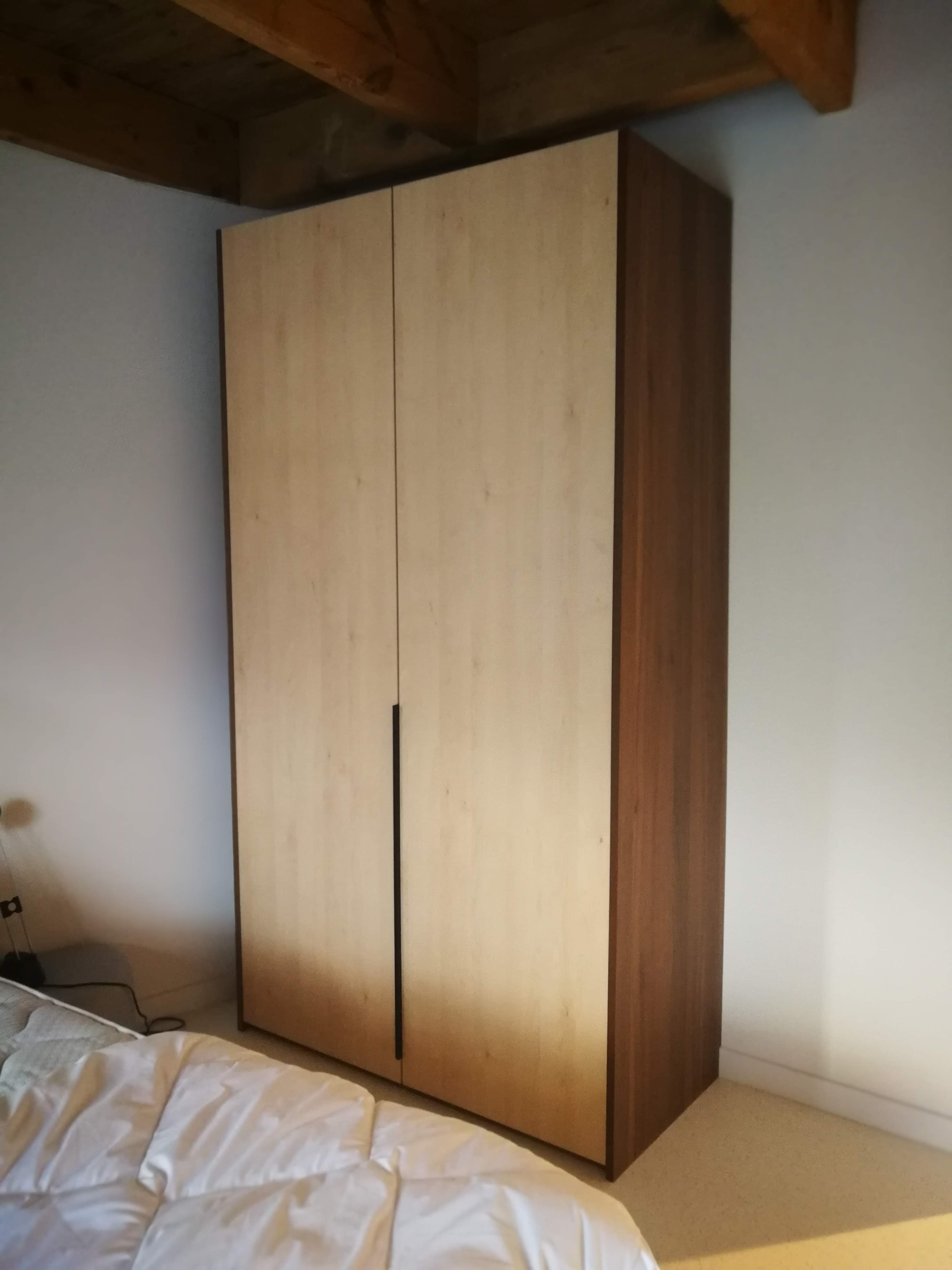 Armario alto em madeira clara com laterais em tom mais escuro, vigas a vista e cama branca