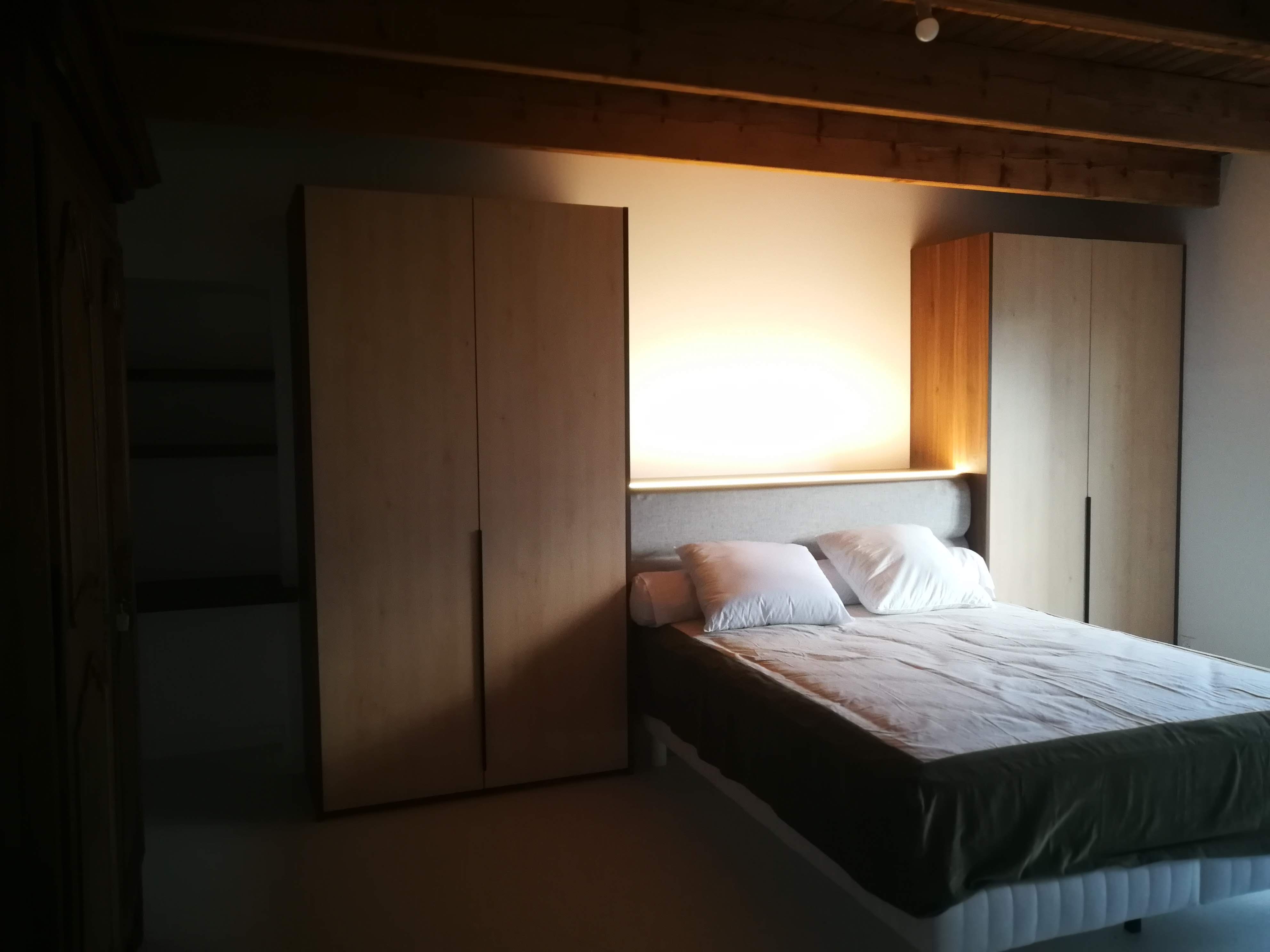 Suite com cama entre armarios em madeira clara, cabeceira em tecido cinza e painel em nogueira com LED